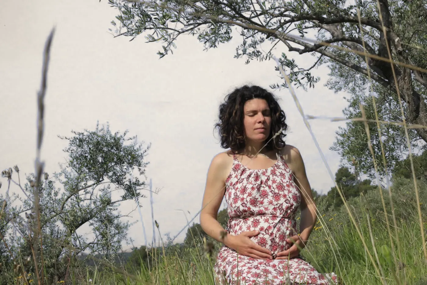 Une femme enceinte assise dans les herbes hautes, les mains posées sur son ventre.