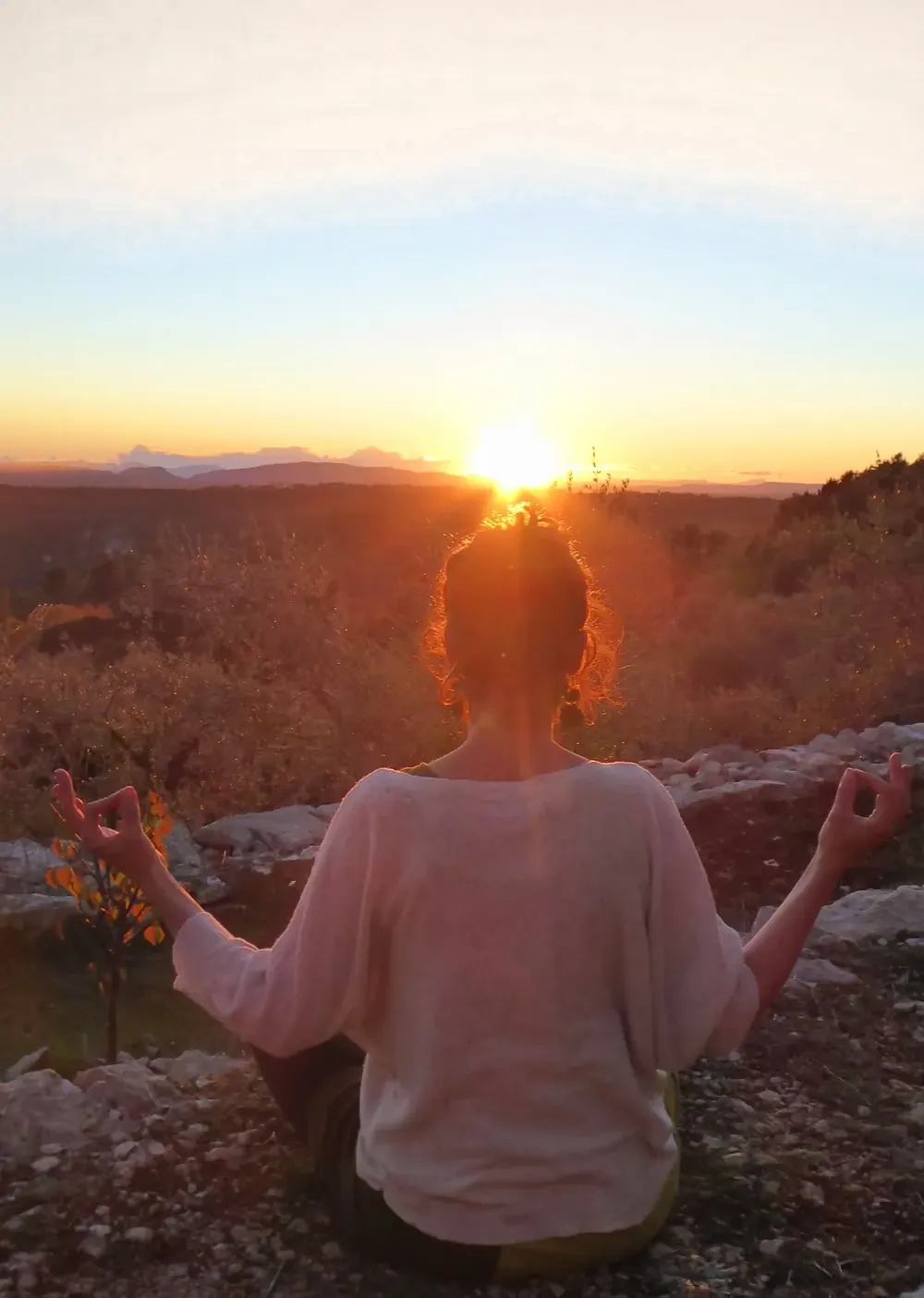 Une femme assise face au soleil couchant, en méditation, dans les collines.