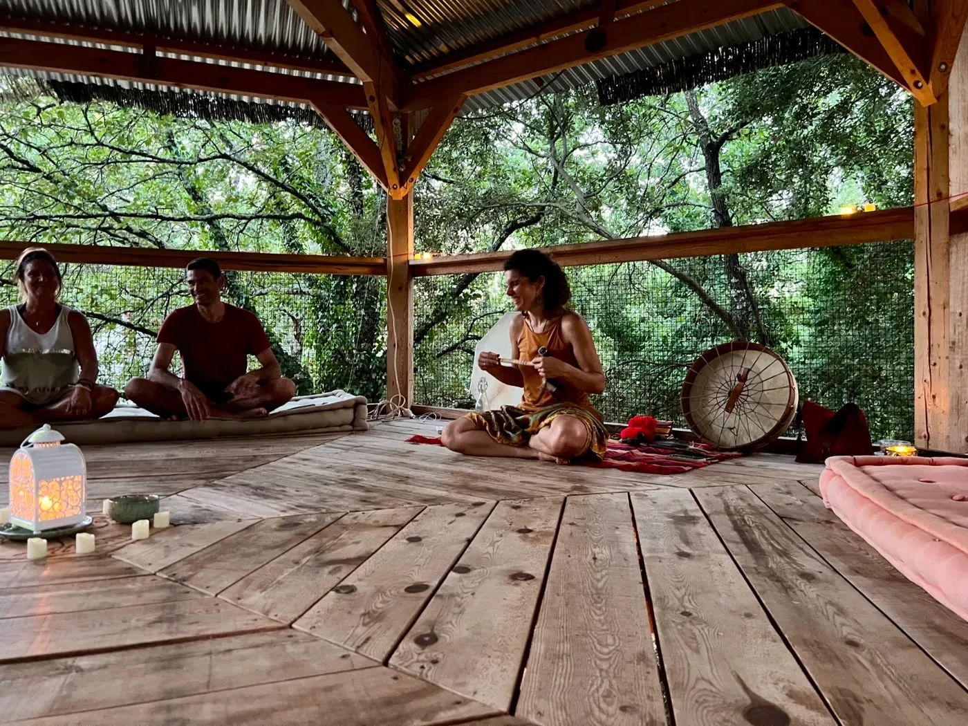 Sous un abri en bois au milieu des arbres, Sandrine joue du tambour devant des participants assis.